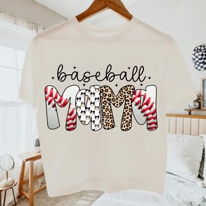 May include: Cream-colored t-shirt with the words "baseball mom" in a playful font. The word "baseball" is in black script, and "MOM" features baseball stitching, leopard print, and bow patterns. A fun, casual top for baseball fans.
