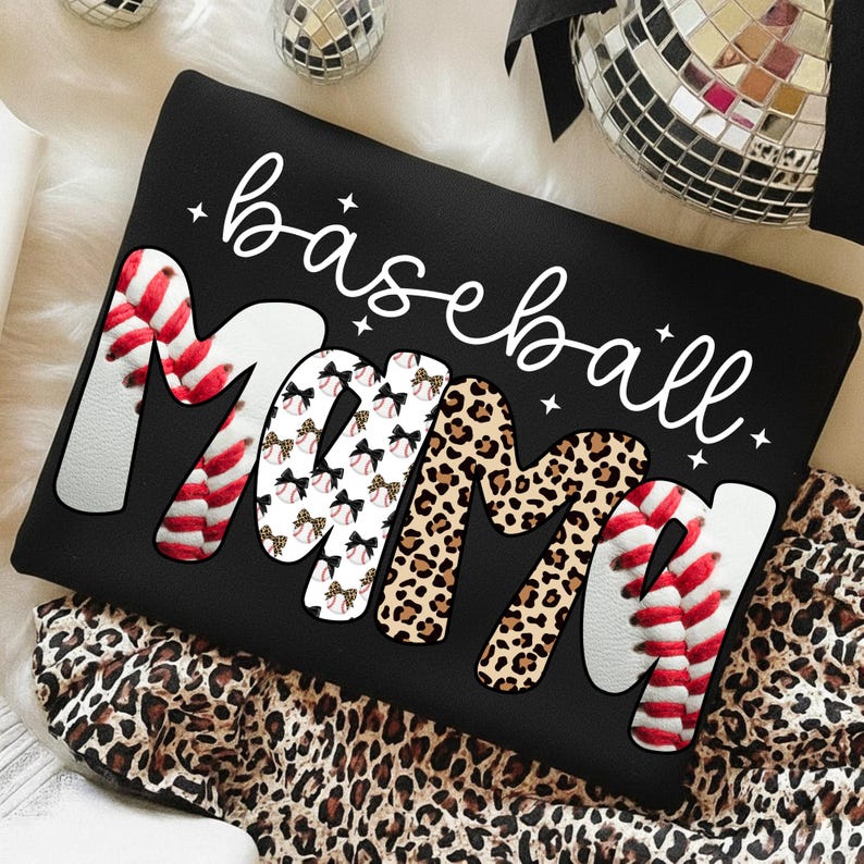 May include: Black sweatshirt with the words "baseball Mama" in white script. The word "Mama" is decorated with baseball stitching, bows, and leopard print. The sweatshirt is on a leopard print fabric.
