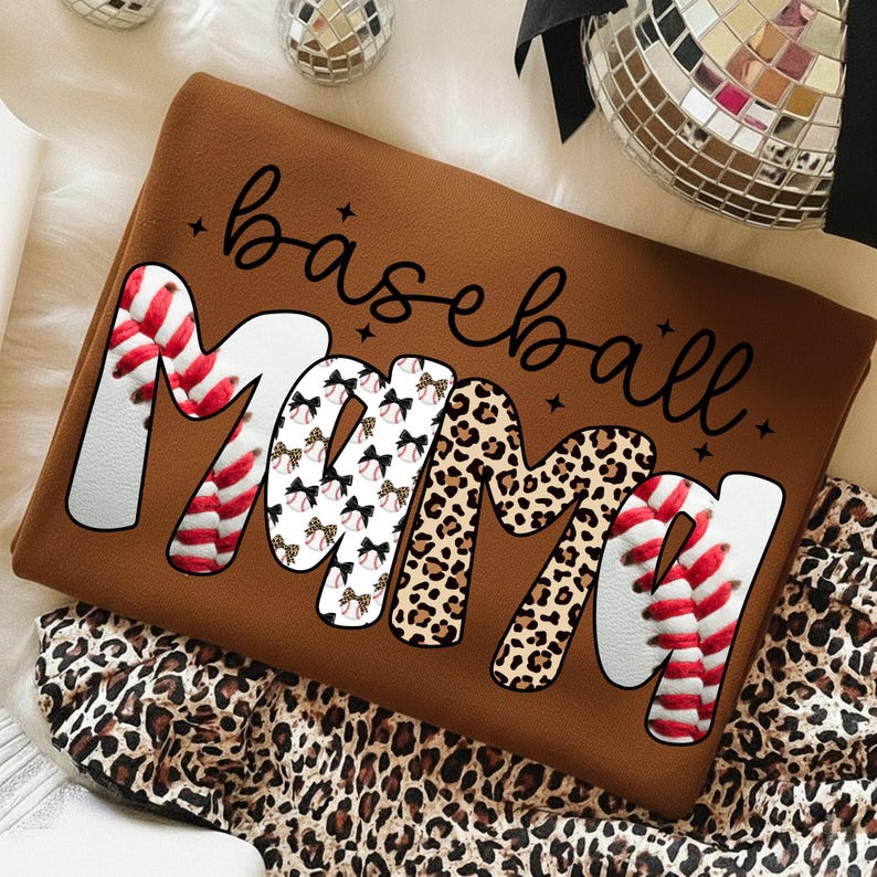 May include: Brown sweatshirt with the words "baseball Mama" in a mix of fonts and patterns. The "M"s are decorated with baseball stitching and leopard print. The sweatshirt is folded on a leopard print fabric.