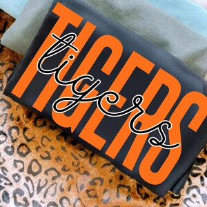 May include: A folded black t-shirt with the word "TIGERS" in large orange block letters. The word "tigers" is also written in a cursive font in white with a black outline. The shirt is on a leopard print surface.