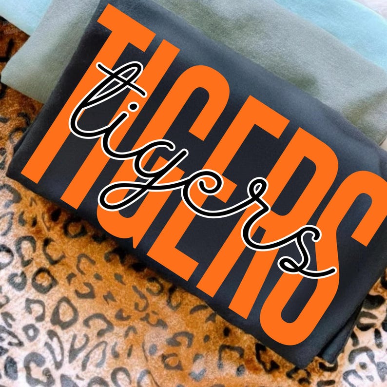 May include: A folded black t-shirt with the word "TIGERS" in large orange block letters. The word "tigers" is also written in a cursive font in white with a black outline. The shirt is on a leopard print surface.