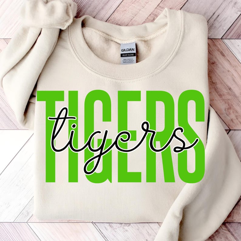 May include: Cream-colored sweatshirt with the word "TIGERS" in large, bright green block letters, with the word "tigers" in a black cursive font overlaying the green letters. The sweatshirt is folded.