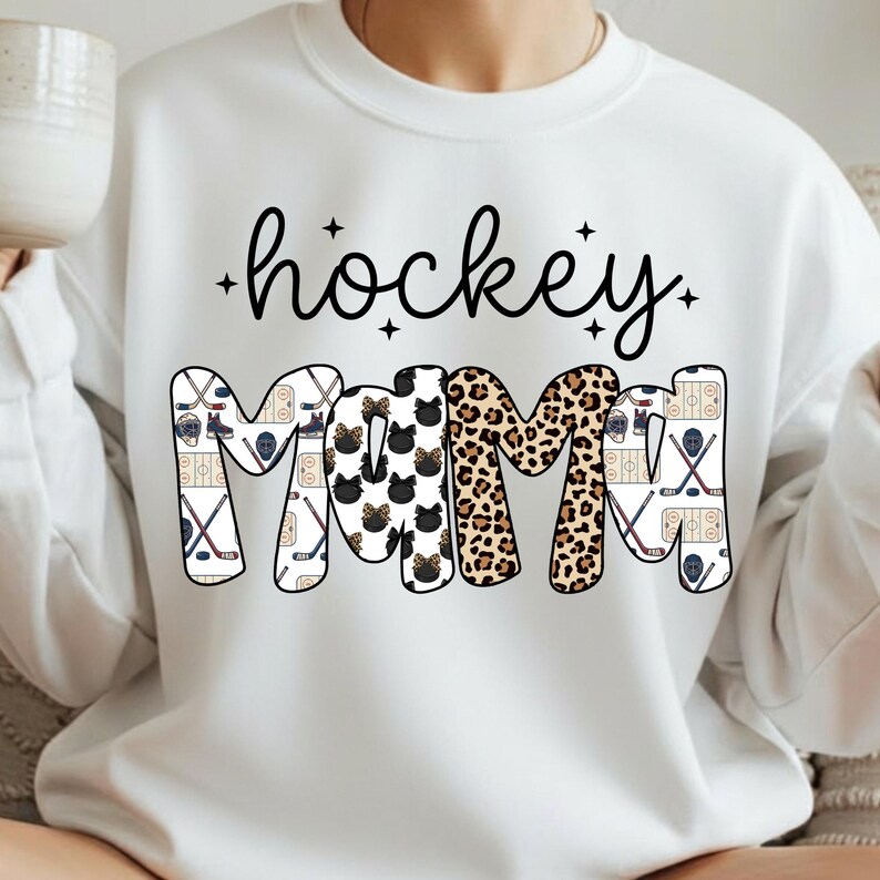 May include: White sweatshirt with the words "hockey mom" in black script and block letters. The letters "M" are decorated with black cat faces and leopard print. The other letters are decorated with hockey sticks and ice rinks.