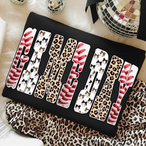 May include: Black sweatshirt with the words "GAME DAY" in a bold, playful font. The letters are decorated with red and white stripes, leopard print, and baseballs with bows. The sweatshirt is folded on a leopard print fabric.