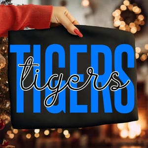 May include: Black fabric with the word "TIGERS" in large blue block letters, with the word "tigers" in a cursive font overlaying the blue letters. The fabric is held by a person with a red sleeve and red nail polish.