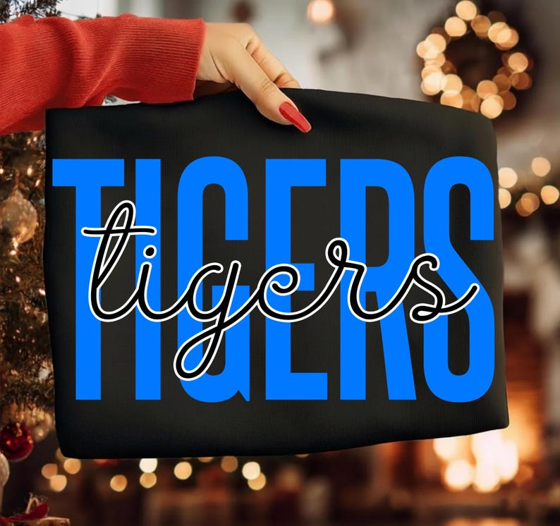 May include: Black fabric with the word "TIGERS" in large blue block letters, with the word "tigers" in a cursive font overlaying the blue letters. The fabric is held by a person with a red sleeve and red nail polish.