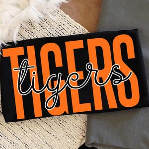 May include: Black t-shirt with the word "TIGERS" in large orange block letters. The word "tigers" is written in a cursive font in white with a black outline. The shirt is folded.