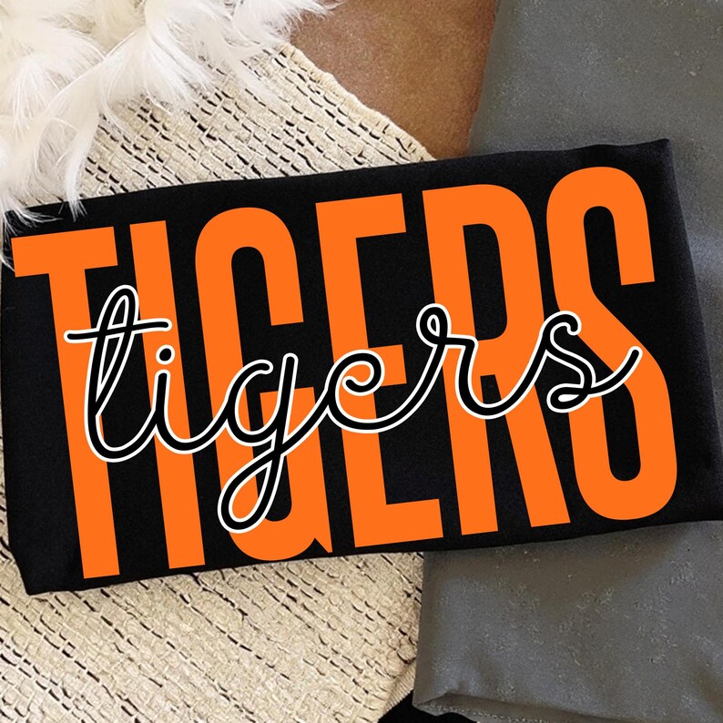May include: Black t-shirt with the word "TIGERS" in large orange block letters. The word "tigers" is written in a cursive font in white with a black outline. The shirt is folded.