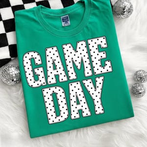 May include: A teal t-shirt with the words "GAME DAY" in large white letters filled with black polka dots. The shirt is displayed on a white fluffy surface with silver disco balls and a black and white checkered pattern.