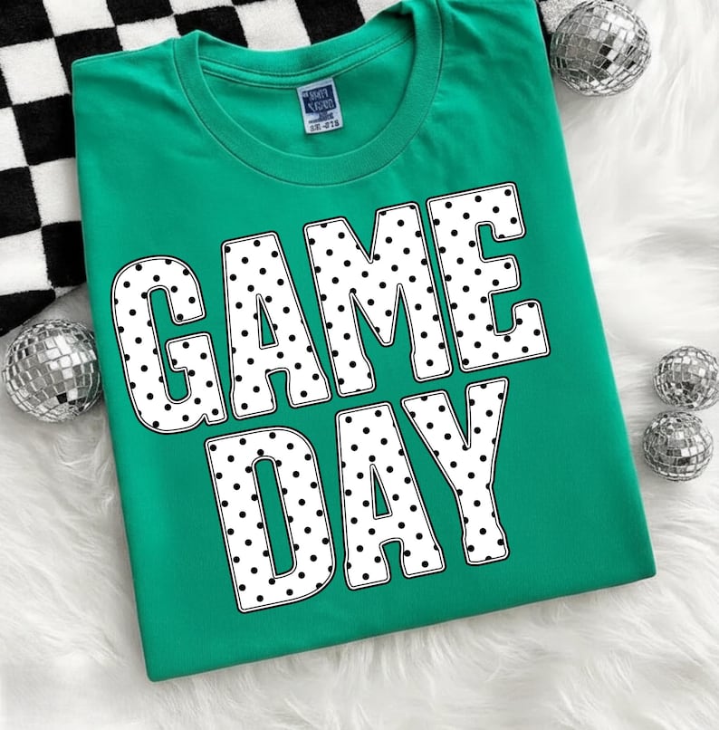 May include: A teal t-shirt with the words "GAME DAY" in large white letters filled with black polka dots. The shirt is displayed on a white fluffy surface with silver disco balls and a black and white checkered pattern.