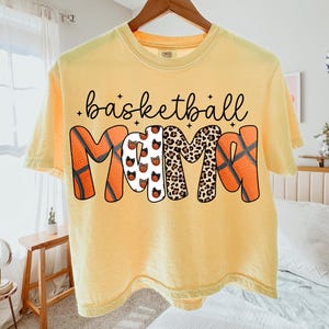 May include: Yellow t-shirt with the words "basketball MOM" in black script and decorative lettering. The "M"s are designed with basketballs and leopard print. The shirt is hanging on a wooden hanger.