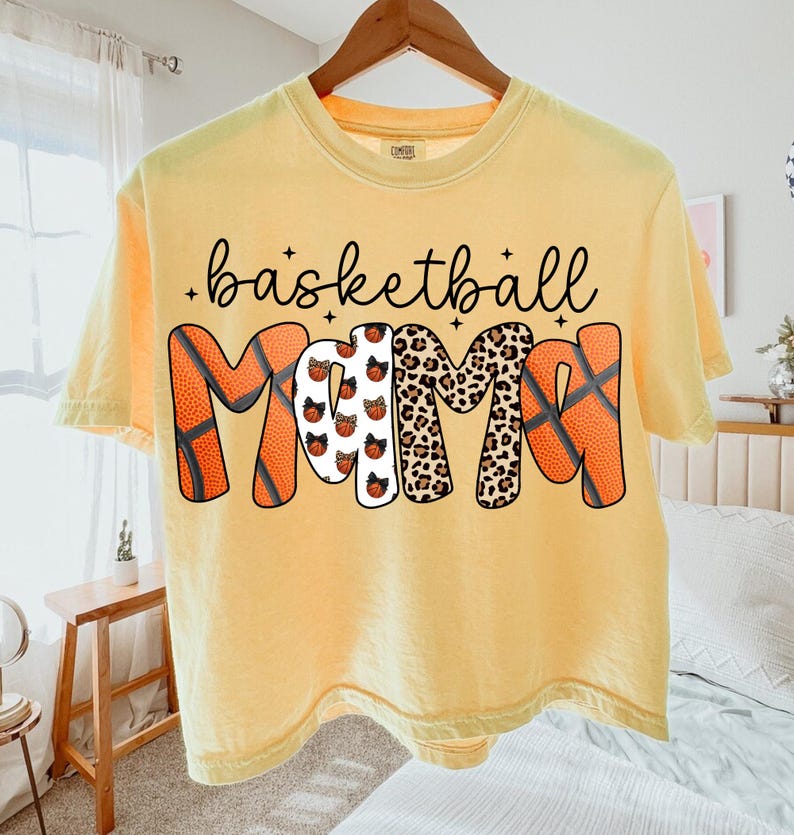 May include: Yellow t-shirt with the words "basketball MOM" in black script and decorative lettering. The "M"s are designed with basketballs and leopard print. The shirt is hanging on a wooden hanger.