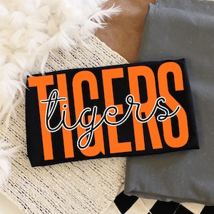 May include: Black rectangular item with the word "TIGERS" in large orange block letters and the word "tigers" in a cursive font. The item is resting on a textured surface with white feathers and gray fabric.