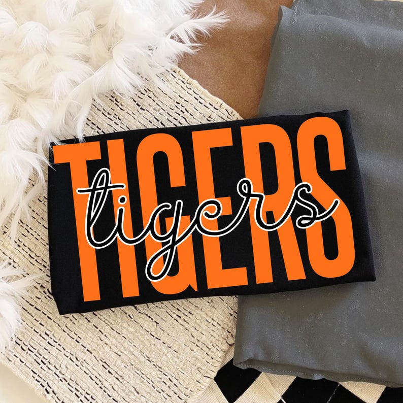 May include: Black rectangular item with the word "TIGERS" in large orange block letters and the word "tigers" in a cursive font. The item is resting on a textured surface with white feathers and gray fabric.