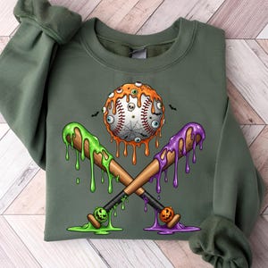 K&ouml;nnte beinhalten: Gr&uuml;nes Sweatshirt mit einem Halloween-Baseball-Design. Der Baseball ist mit Aug&auml;pfeln, einem Totenkopf und einem Spinnennetz verziert. Zwei gekreuzte Baseballschl&auml;ger mit gr&uuml;nem und lila tropfendem Schleim vervollst&auml;ndigen das gruselige Design.
