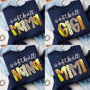 以下が含まれることがあります： 紺色のスウェットシャツ。"softball MOM", "softball GIGI", "softball NANA", "softball MIMI"の文字が黄色、ヒョウ柄、ソフトボールのデザインでプリントされています。スウェットシャツは青いジーンズの背景に表示されています。