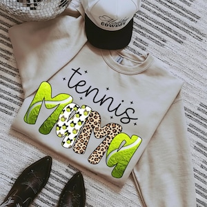 K&ouml;nnte beinhalten: Ein beiges Sweatshirt mit den Worten "tennis Mama" in einer Mischung aus Schriftarten und Mustern, darunter Tennisballgrafiken und Leopardenmuster. Eine wei&szlig;-schwarze Trucker-M&uuml;tze und schwarze Stiefel sind ebenfalls im Bild.