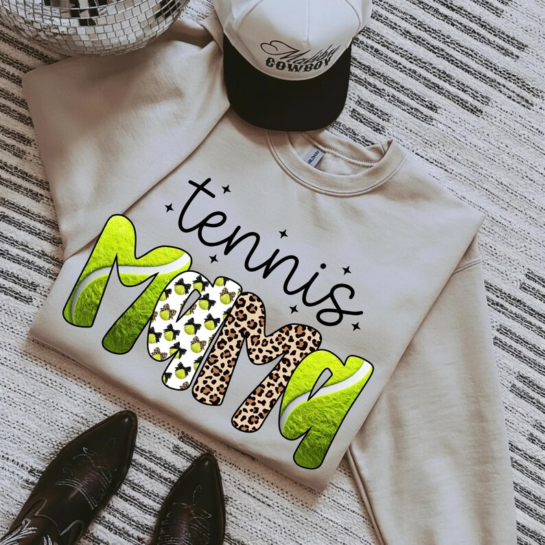 K&ouml;nnte beinhalten: Ein beiges Sweatshirt mit den Worten "tennis Mama" in einer Mischung aus Schriftarten und Mustern, darunter Tennisballgrafiken und Leopardenmuster. Eine wei&szlig;-schwarze Trucker-M&uuml;tze und schwarze Stiefel sind ebenfalls im Bild.