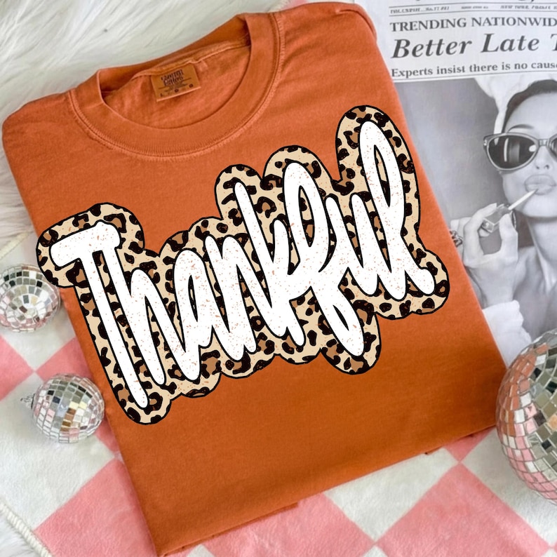 Peut inclure: T-shirt orange avec le mot "Thankful" en blanc, bord&eacute; d'un motif l&eacute;opard. Le t-shirt est pli&eacute; sur une surface &agrave; carreaux roses et blancs, avec des boules &agrave; facettes d&eacute;coratives.