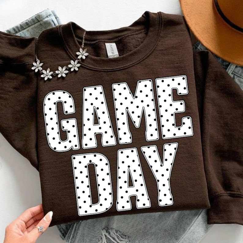 May include: A brown sweatshirt with the words "GAME DAY" in large white letters with black polka dots. A silver necklace with flower-shaped charms is draped on the sweatshirt. The sweatshirt is on top of a pair of blue jeans.