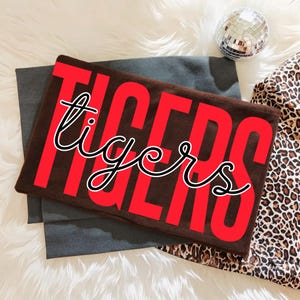 May include: A brown fabric item with the word "TIGERS" in large red letters, with the word "tigers" in a cursive font overlaying the red letters. The item is on a gray fabric, with a disco ball and leopard print fabric in the background.