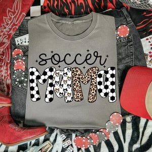 May include: A gray t-shirt with the words "soccer mama" in black script. The word "mama" is designed with soccer ball patterns, leopard print, and soccer ball dog faces. The shirt is surrounded by denim jeans, a red hat, and poker chips.