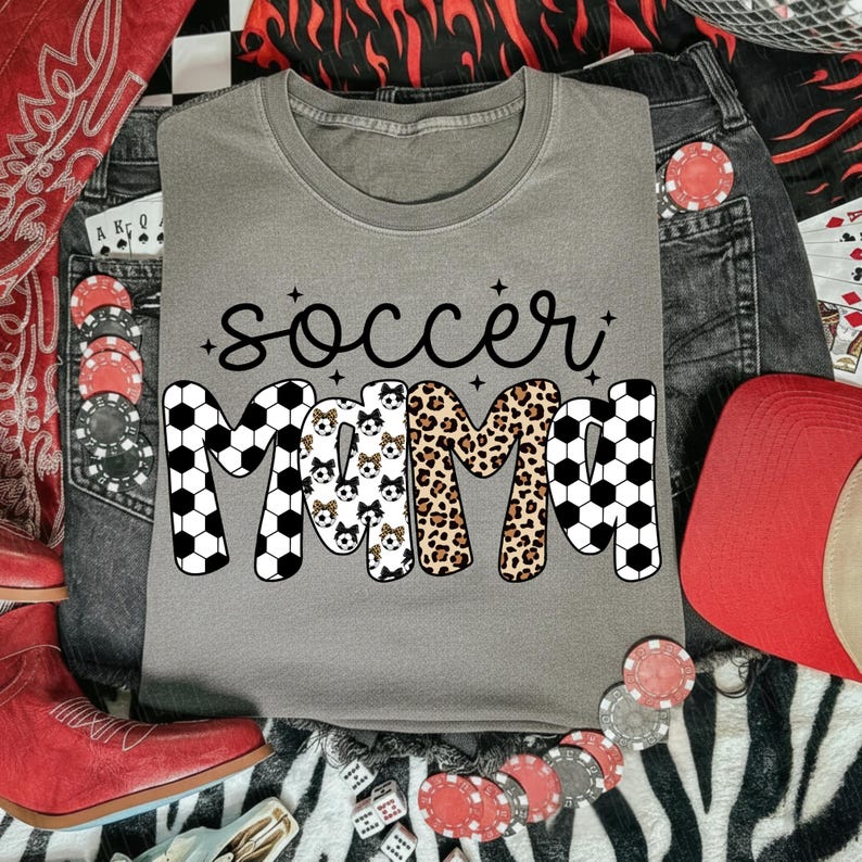 May include: A gray t-shirt with the words "soccer mama" in black script. The word "mama" is designed with soccer ball patterns, leopard print, and soccer ball dog faces. The shirt is surrounded by denim jeans, a red hat, and poker chips.