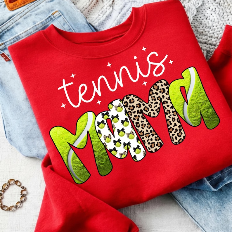 K&ouml;nnte beinhalten: Ein rotes Sweatshirt mit dem wei&szlig;en Schriftzug "tennis mama". Das Wort "mama" ist mit Tennisball- und Leopardenmuster verziert. Das Sweatshirt ist auf einer hellen Oberfl&auml;che gefaltet, mit einer Jeans sichtbar.