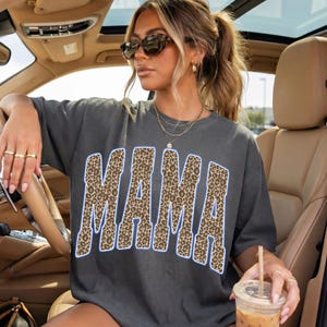 May include: A charcoal gray short-sleeve t-shirt featuring the word MAMA in large, arched block letters with a brown and black leopard print pattern and a thin blue outline. The shirt has a crew neckline and a relaxed, loose-fitting silhouette.