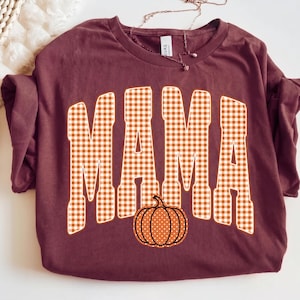 May include: A maroon short-sleeved t-shirt featuring the word MAMA in large, block-style letters with an orange and white gingham check pattern. Below the text is a small, round orange pumpkin graphic with white polka dots and a black outline.