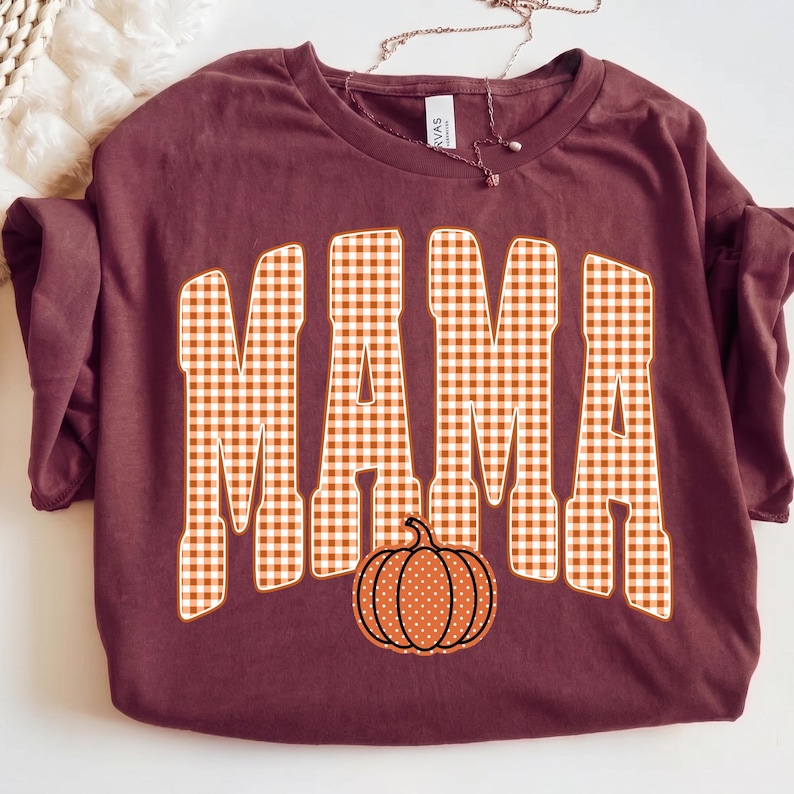 May include: A maroon short-sleeved t-shirt featuring the word MAMA in large, block-style letters with an orange and white gingham check pattern. Below the text is a small, round orange pumpkin graphic with white polka dots and a black outline.