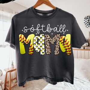May include: Dark gray t-shirt with the words "softball mama" in white script. The word "mama" is designed with a softball pattern, leopard print, and softball stitching. A casual tee for softball fans.