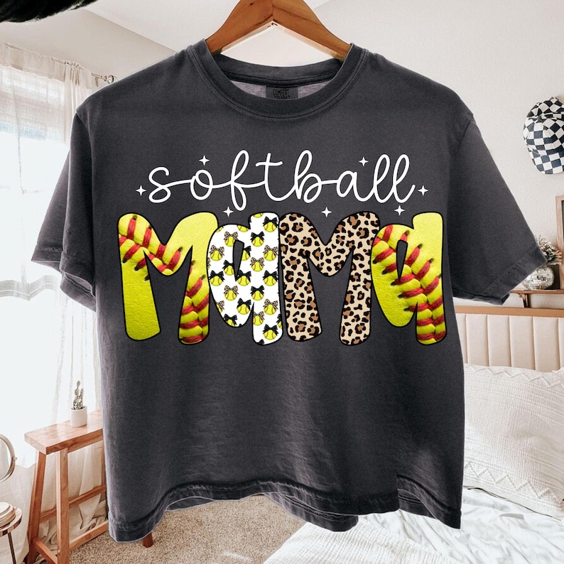 May include: Dark gray t-shirt with the words "softball mama" in white script. The word "mama" is designed with a softball pattern, leopard print, and softball stitching. A casual tee for softball fans.
