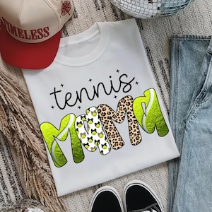 K&ouml;nnte beinhalten: Wei&szlig;es T-Shirt mit den Worten "tennis mama" in schwarzer Schrift und fetten, farbenfrohen Buchstaben. Das "M" ist mit Tennisb&auml;llen und Schleifen verziert, und das andere "M" ist im Leopardenmuster. Eine rote und beige Baseballkappe ist im Hintergrund.
