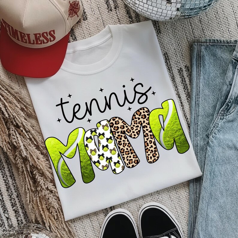 K&ouml;nnte beinhalten: Wei&szlig;es T-Shirt mit den Worten "tennis mama" in schwarzer Schrift und fetten, farbenfrohen Buchstaben. Das "M" ist mit Tennisb&auml;llen und Schleifen verziert, und das andere "M" ist im Leopardenmuster. Eine rote und beige Baseballkappe ist im Hintergrund.