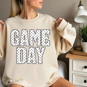 May include: Cream-colored sweatshirt with the words "GAME DAY" in large, black-outlined letters filled with white polka dots. The sweatshirt has long sleeves and a relaxed fit, perfect for casual wear.