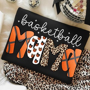 May include: Black sweatshirt with the words "basketball Mama" in white script. The word "Mama" is designed with basketball textures, leopard print, and basketball graphics. The sweatshirt is on a leopard print fabric.