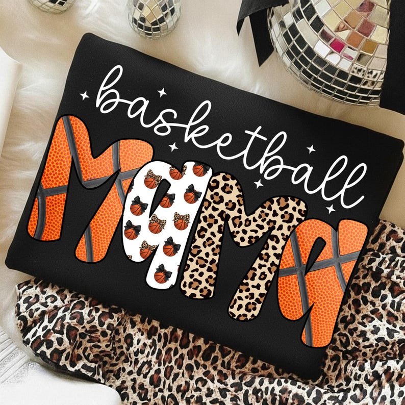 May include: Black sweatshirt with the words "basketball Mama" in white script. The word "Mama" is designed with basketball textures, leopard print, and basketball graphics. The sweatshirt is on a leopard print fabric.