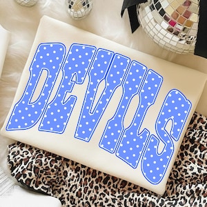 May include: A cream-colored sweatshirt featuring the word DEVILS in large, bold, blue letters with a white polka dot pattern. The text has a thick blue outline and is centered on the front of the garment, which is folded neatly.