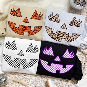 May include: Four sweatshirts with jack-o'-lantern faces. The faces have triangle eyes and a smiling mouth. The faces are in different patterns: leopard print, black and white checkerboard, and purple and white checkerboard. The sweatshirts are in beige, gray, white, and black.