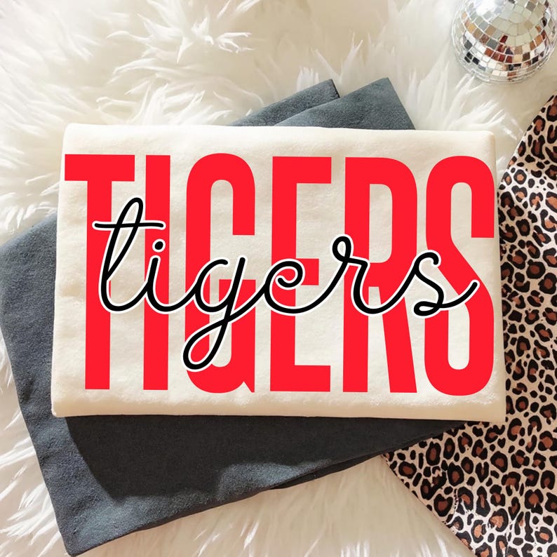 May include: Cream-colored t-shirt with the word "TIGERS" in large red block letters, and the word "tigers" in a cursive font overlaying the red letters. The shirt is folded on top of a dark gray fabric, with a leopard print fabric and a disco ball in the background.
