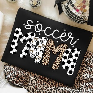 May include: Black sweatshirt with the words "Soccer Mama" in white script and bold letters. The letters "M" are decorated with soccer balls and leopard print. The sweatshirt is on a leopard print fabric.
