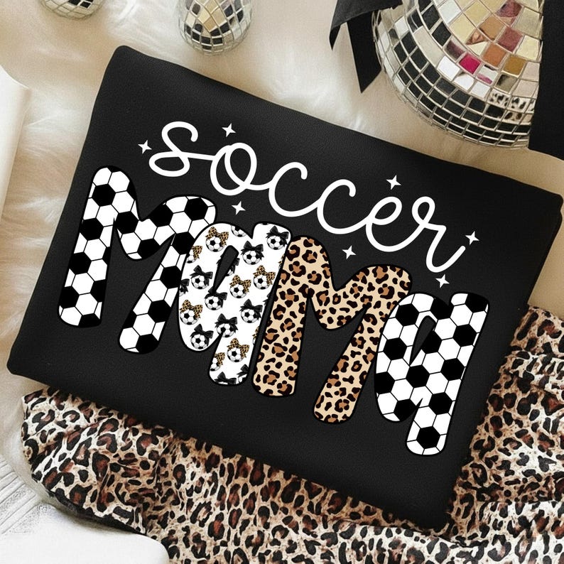 May include: Black sweatshirt with the words "Soccer Mama" in white script and bold letters. The letters "M" are decorated with soccer balls and leopard print. The sweatshirt is on a leopard print fabric.