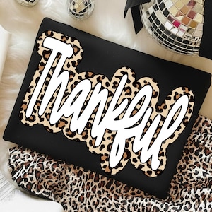 Peut inclure: Sweat-shirt noir avec le mot "Thankful" en &eacute;criture blanche bord&eacute; d'un motif l&eacute;opard. Le v&ecirc;tement est pli&eacute; sur une surface avec un motif l&eacute;opard. Des boules disco sont visibles en arri&egrave;re-plan.