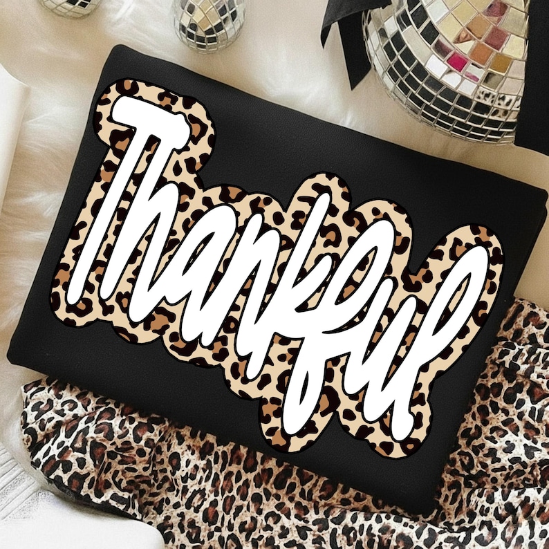 Peut inclure: Sweat-shirt noir avec le mot "Thankful" en &eacute;criture blanche bord&eacute; d'un motif l&eacute;opard. Le v&ecirc;tement est pli&eacute; sur une surface avec un motif l&eacute;opard. Des boules disco sont visibles en arri&egrave;re-plan.