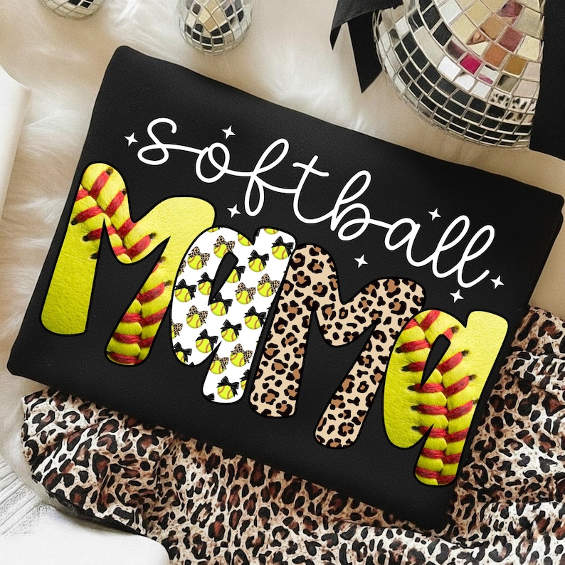 May include: Black sweatshirt with the words "softball mama" in white script. The word "Mama" is designed with a softball, leopard print, and softball pattern. The sweatshirt is on a leopard print fabric.