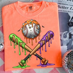K&ouml;nnte beinhalten: Orangefarbenes T-Shirt mit einem Halloween-Baseball-Design. Der Baseball hat tropfenden orangen Schleim, Augen und ein Spinnennetz. Zwei gekreuzte Schl&auml;ger mit gr&uuml;nem und lila tropfendem Schleim.