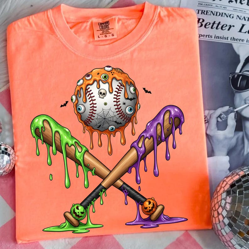 K&ouml;nnte beinhalten: Orangefarbenes T-Shirt mit einem Halloween-Baseball-Design. Der Baseball hat tropfenden orangen Schleim, Augen und ein Spinnennetz. Zwei gekreuzte Schl&auml;ger mit gr&uuml;nem und lila tropfendem Schleim.