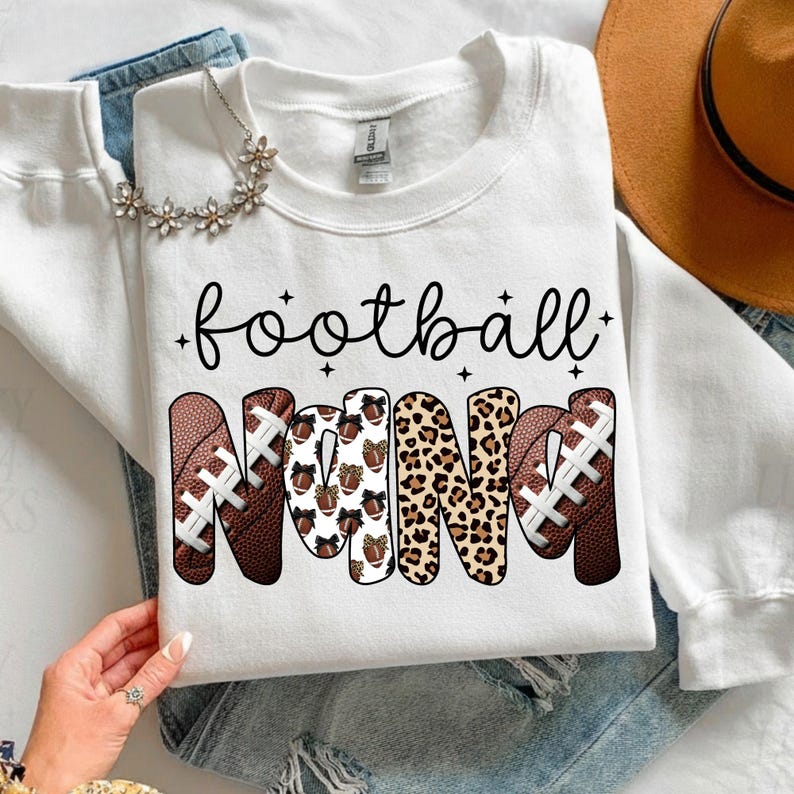May include: White sweatshirt with the words "football NANA" in black script and football-themed letters. The letters are designed with footballs, leopard print, and football patterns. A floral necklace and a brown hat are also visible.