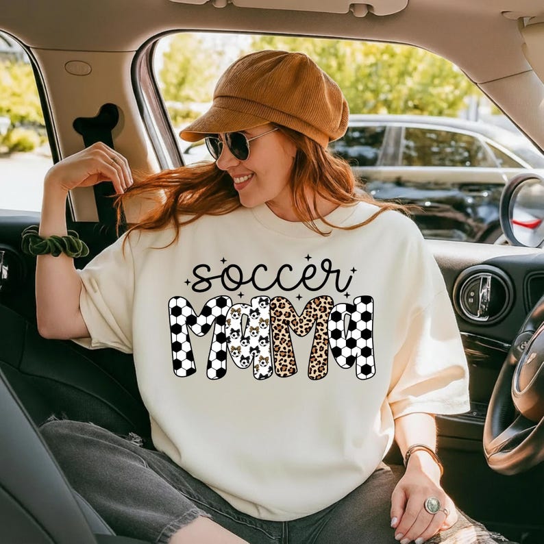 May include: Cream-colored sweatshirt with the text "soccer mama" in a mix of black and white soccer ball patterns, leopard print, and dog silhouettes. The shirt is worn by a person in a car.